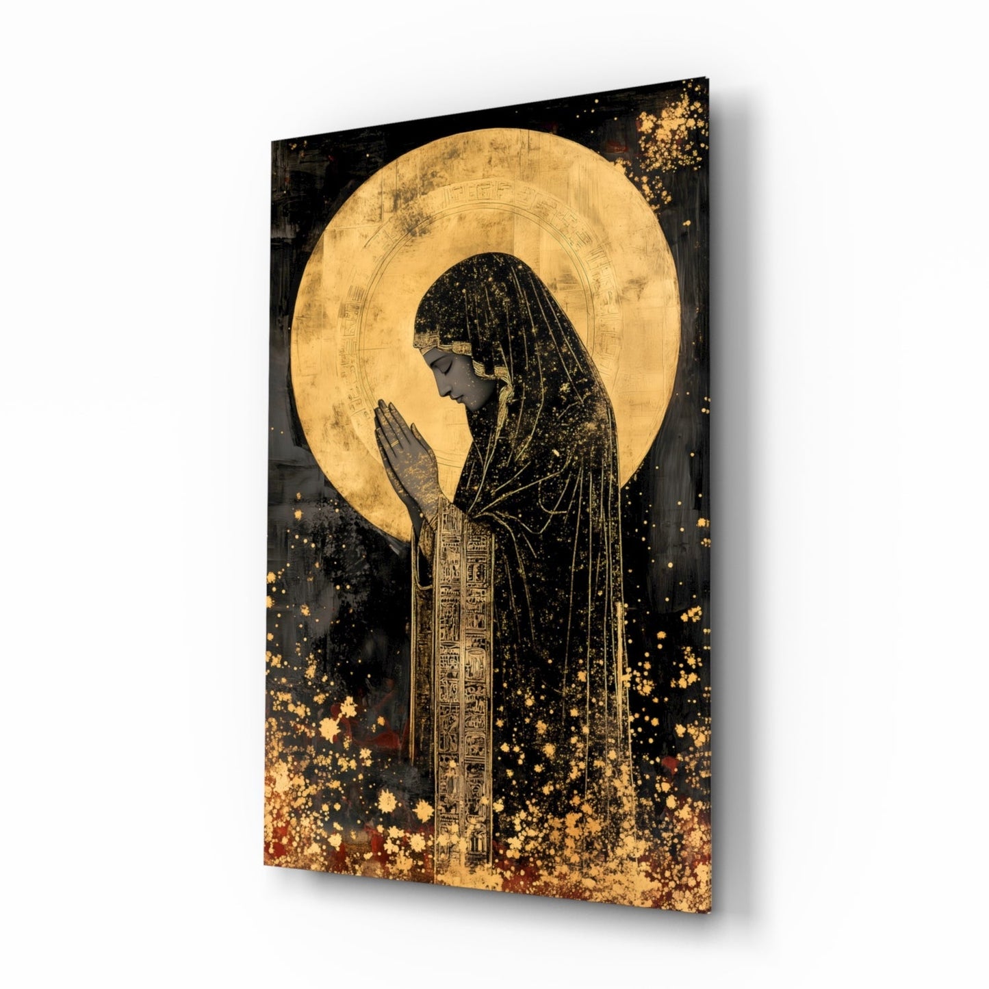 Angled view of glass wall art depicting a praying figure in profile with gold leaf halo against black background and gold splatter effects.