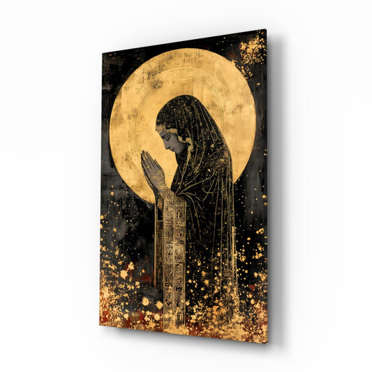 Angled view of glass wall art depicting a praying figure in profile with gold leaf halo against black background and gold splatter effects.