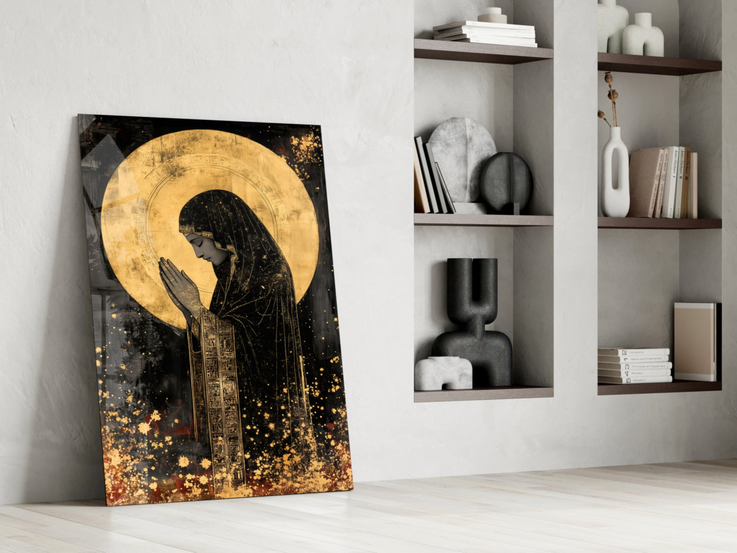 Golden Prayers of Mary glass wall art leaning against white wall beside built-in shelving displaying decorative objects and books.
