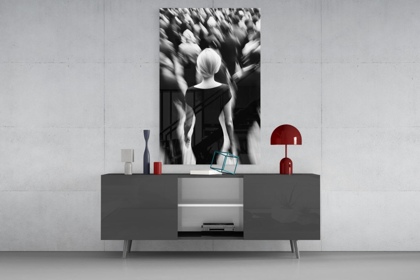 Black and white glass wall art depicting a solitary figure surrounded by blurred crowd, displayed above gray console table with red lamp and accessories.