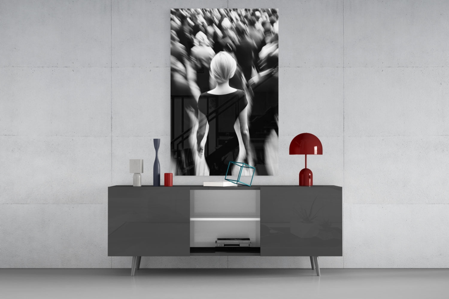 Black and white glass wall art depicting a solitary figure surrounded by blurred crowd, displayed above gray console table with red lamp and accessories.