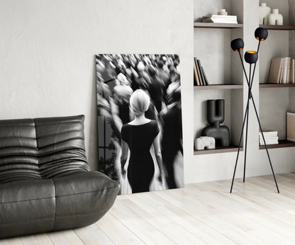 Black and white glass wall art depicting a solitary figure viewed from behind surrounded by blurred crowd, displayed in modern living room with leather sofa and shelving.