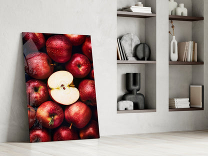 Glass wall art leaning against white wall displays red apples with one sliced in half, next to built-in shelving with decorative objects.