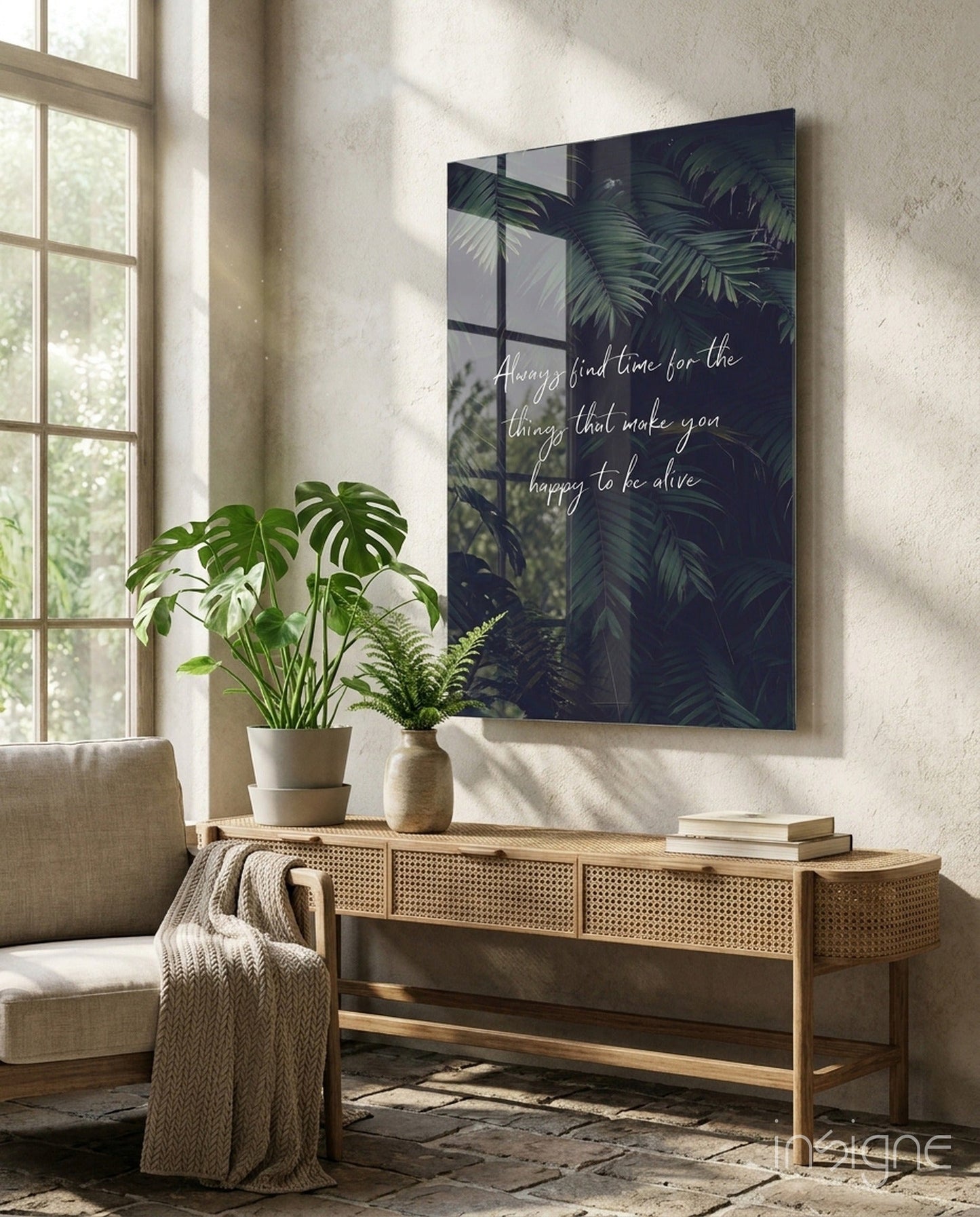 Dark glass wall art with white cursive text and tropical foliage displayed above wooden console table with potted plants.