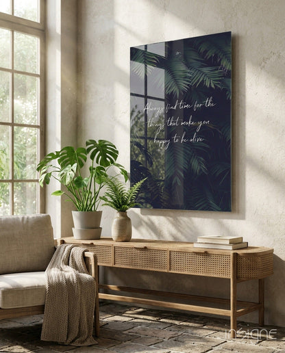 Dark glass wall art with white cursive text and tropical foliage displayed above wooden console table with potted plants.