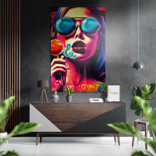 Vibrant pop art portrait of woman wearing sunglasses and holding orange lollipop, displayed on gray wall above wooden dresser.