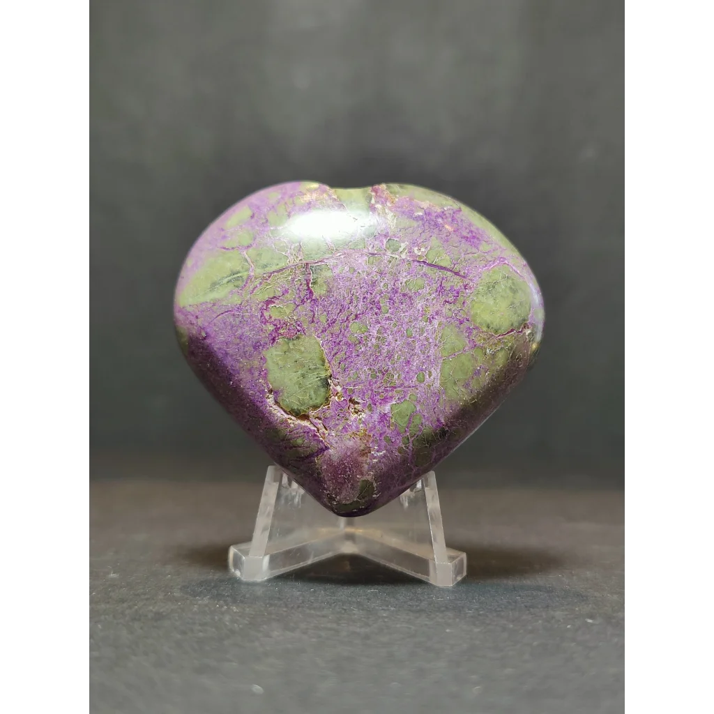 A polished heart-shaped atlantisite mineral specimen with vibrant purple and green marbled pattern on a clear stand