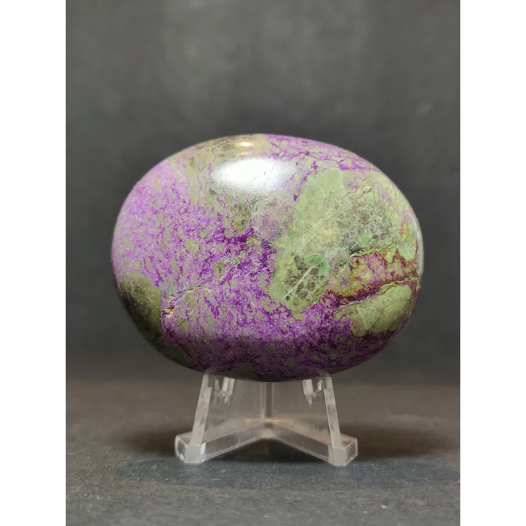 A polished oval-shaped purple and green atlantisite mineral heart and palmstone sculpture displayed on a clear acrylic stand