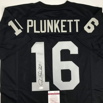 Back side of an autographed black jim plunkett football jersey with white custom lettering and number 16 from a professional sports league