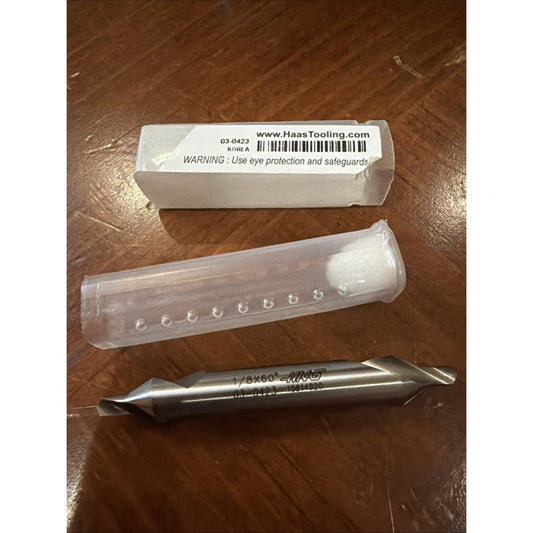 Haas Tooling 1/8" 60° chamfer mill with white box, clear plastic tube, and metal drill bit on brown wooden surface.