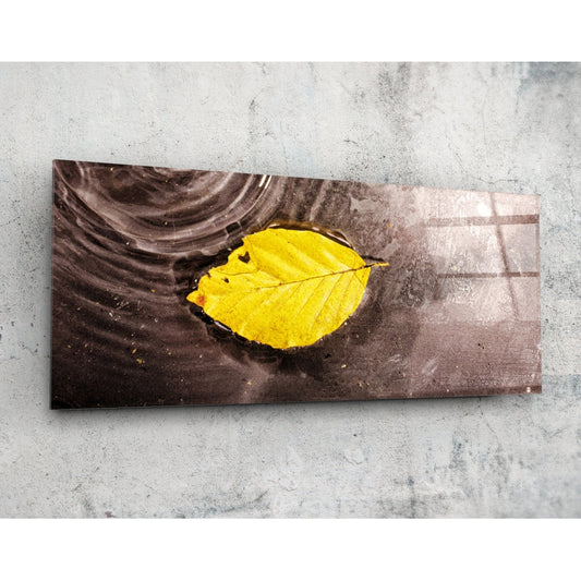 Horizontal rectangular glass art panel displaying a vibrant yellow leaf floating on dark brown water, photographed flat against concrete.