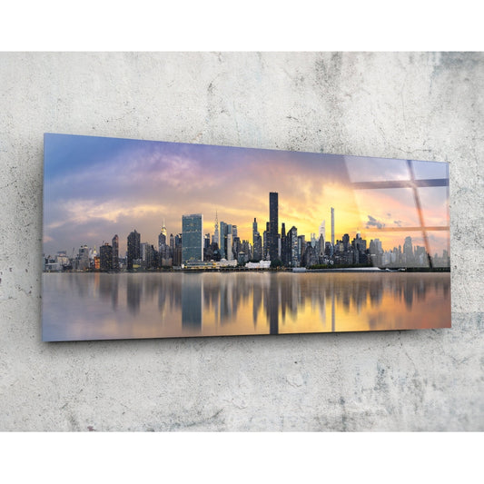 Panoramic glass wall art depicting Manhattan skyline at sunset with golden sky reflected in water, mounted on concrete wall.