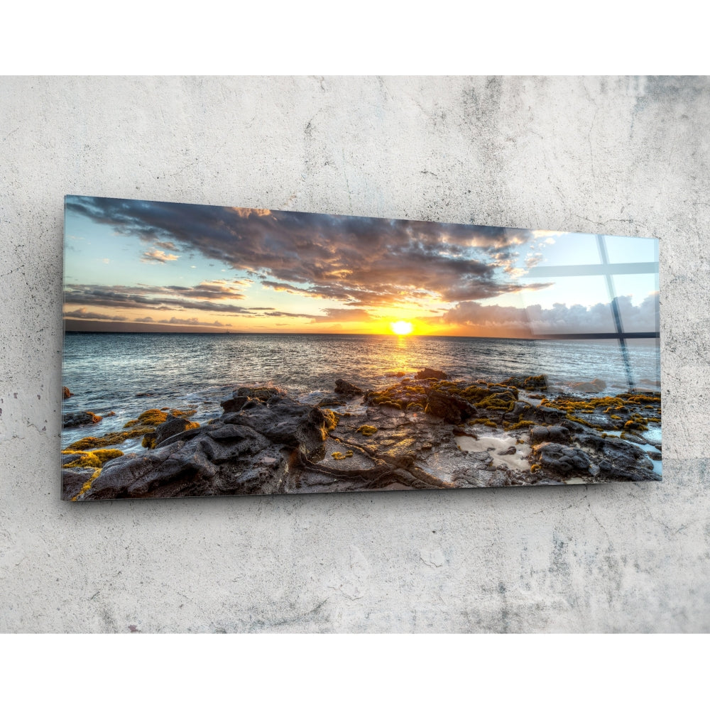 Rectangular glass wall art mounted on concrete showing a coastal sunset with golden sun, rocky shoreline, and dramatic clouds reflected in glossy surface.