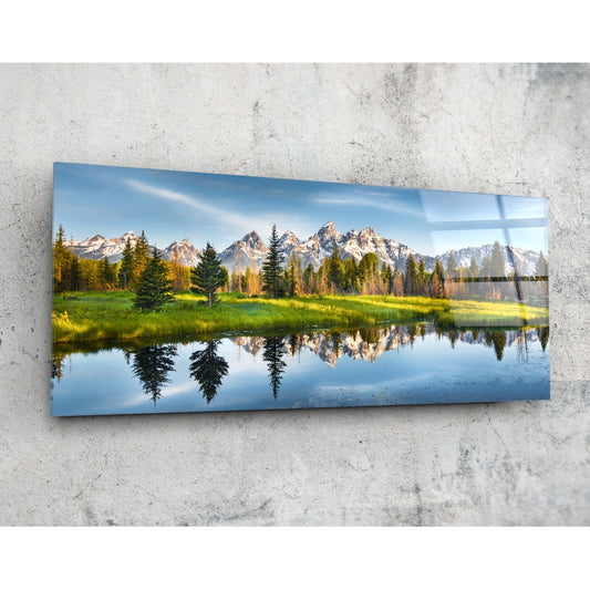 Rectangular glass wall art mounted on concrete displaying mountain landscape with evergreen trees, meadow, and mirrored water reflection under blue sky.