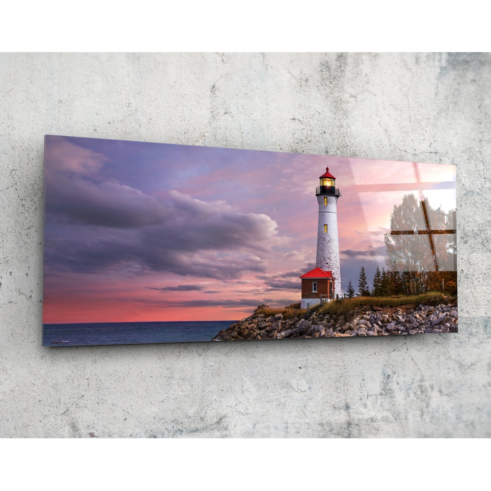 Rectangular glass wall art mounted on concrete displaying a lighthouse with red roof at sunset over rocky shoreline with evergreen trees and wooden cross.
