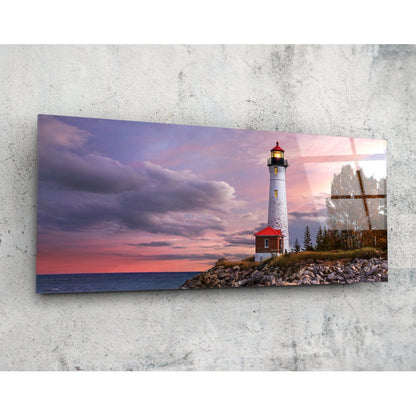 Rectangular glass wall art mounted on concrete displaying a lighthouse with red roof at sunset over rocky shoreline with evergreen trees and wooden cross.