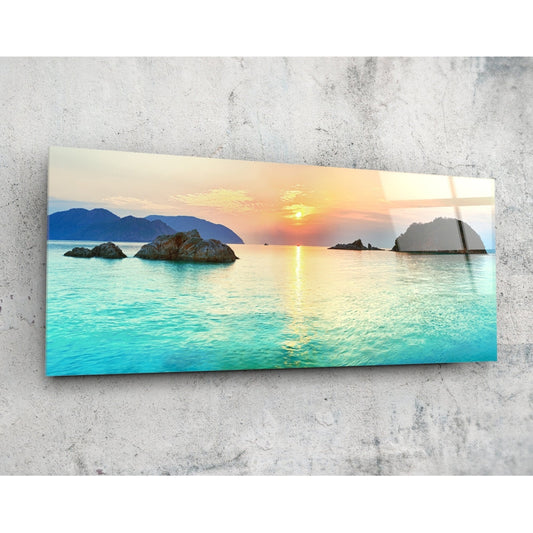 Glass wall art panel mounted on concrete showing turquoise bay with rocky islets at sunset, golden sky reflected in water.