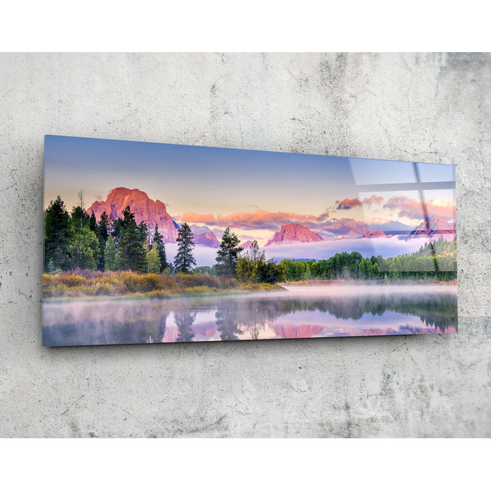 Horizontal glass wall art mounted on concrete displaying a misty sunrise landscape with mountains, forest, and calm lake reflection in pink and blue tones.