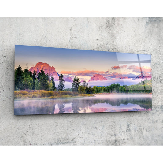 Horizontal glass wall art mounted on concrete displaying a misty sunrise landscape with mountains, forest, and calm lake reflection in pink and blue tones.