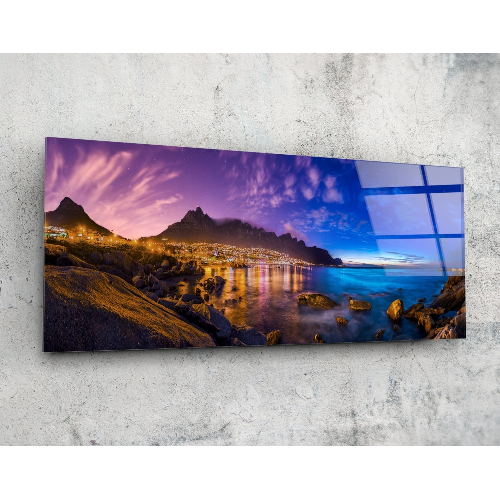 Horizontal glass wall art mounted on concrete showing twilight coastal landscape with illuminated mountainside city, purple sky, and rocky shoreline.