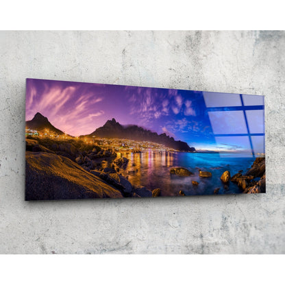Horizontal glass wall art mounted on concrete showing twilight coastal landscape with illuminated mountainside city, purple sky, and rocky shoreline.