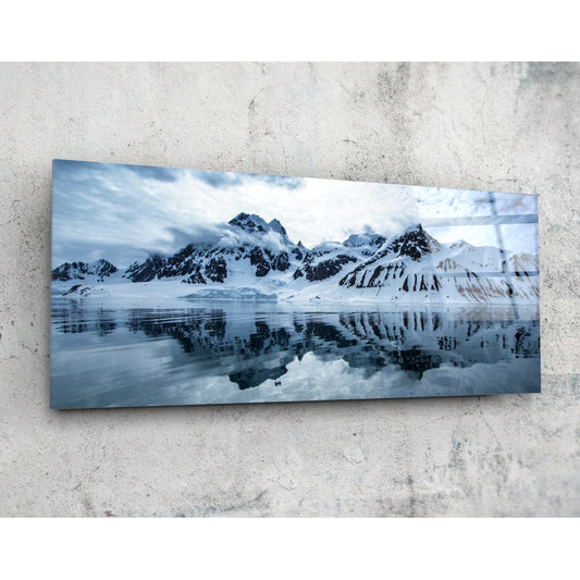 Rectangular glass panel mounted on concrete wall displaying snow-covered mountains reflected in still water beneath cloudy sky.