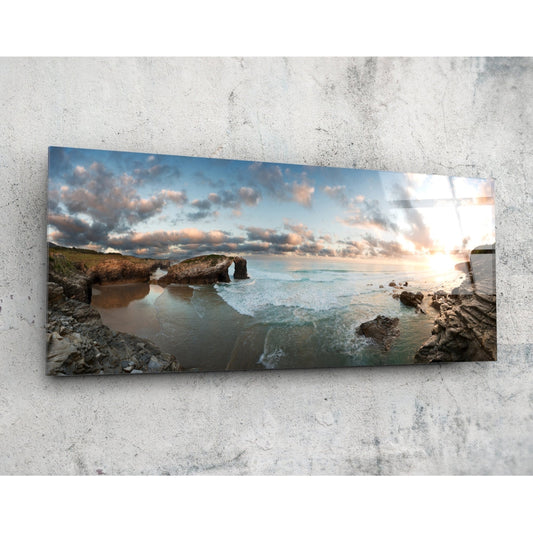 Glass wall art panel mounted on concrete showing a coastal seascape with rocky arch formation, golden sunset, and turquoise waves.