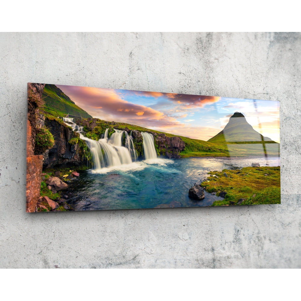 Rectangular glass wall art mounted on concrete showing cascading waterfall, turquoise pool, moss-covered rocks, green mountainside, and sunset sky.