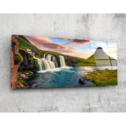 Rectangular glass wall art mounted on concrete showing cascading waterfall, turquoise pool, moss-covered rocks, green mountainside, and sunset sky.