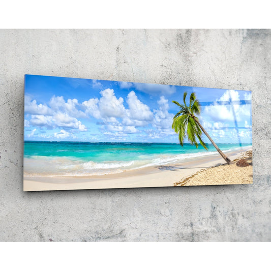 Tropical beach scene glass wall art mounted on concrete wall showing palm tree, sand, turquoise ocean, and blue sky with white clouds.