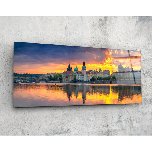 Panoramic glass wall art mounted on concrete displaying Prague skyline with illuminated towers, bridges, and river reflections at golden sunset.