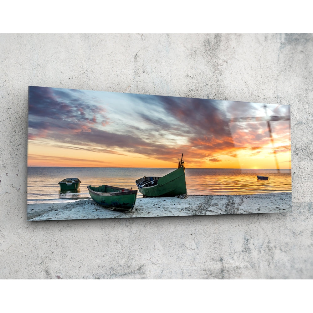 Rectangular glass wall art panel mounted on concrete displaying green fishing boats on a beach at sunset with golden and orange sky.