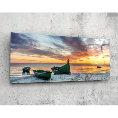 Rectangular glass wall art panel mounted on concrete displaying green fishing boats on a beach at sunset with golden and orange sky.