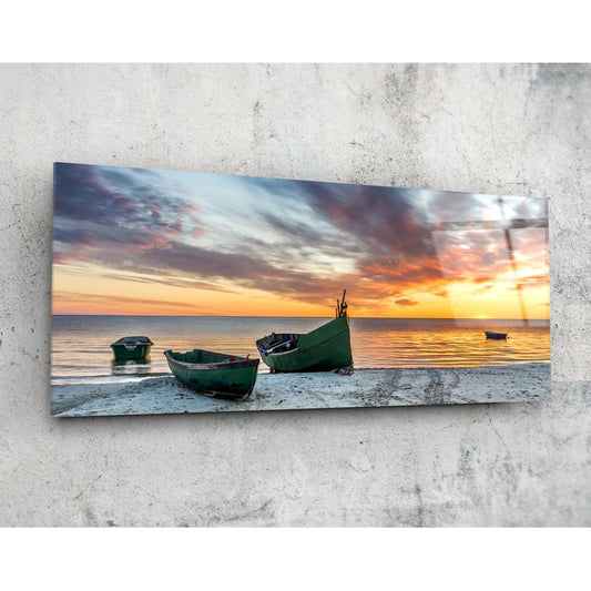 Rectangular glass wall art panel mounted on concrete displaying green fishing boats on a beach at sunset with golden and orange sky.