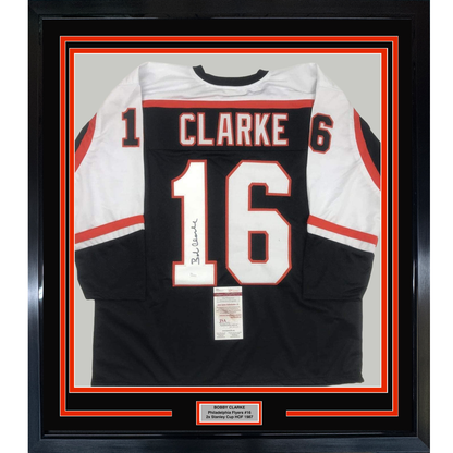 Framed black and white Philadelphia Flyers hockey jersey number 16 with "CLARKE" nameplate, signed, displayed against red and black matting with authentication certificate visible.