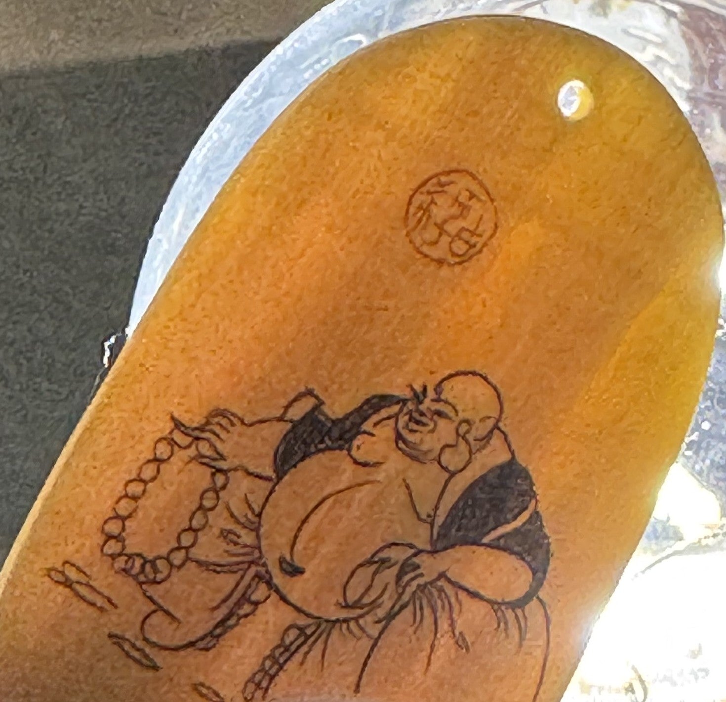 Translucent amber ox horn pendant with carved Budai figure holding beads and a circular seal mark, photographed at angle agai