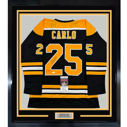 Framed black and gold Boston Bruins jersey number 25 signed by Brandon Carlo, displayed flat against light gray background with authentication certificate visible.