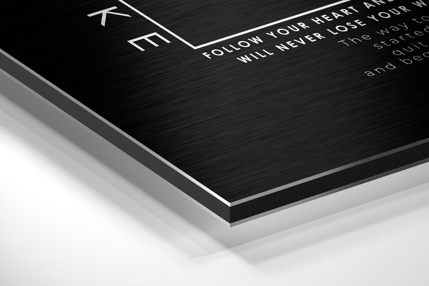 Angled close-up of black brushed aluminum Dibond wall art with white engraved text, showing layered edge detail against white background.