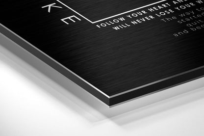 Angled close-up of black brushed aluminum Dibond wall art with white engraved text, showing layered edge detail against white background.