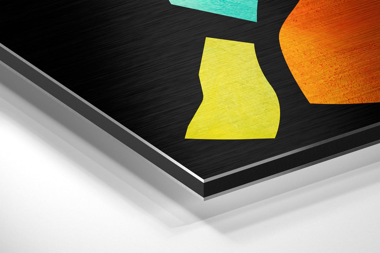 Angled overhead view of brushed aluminum Dibond wall art showing layered edge detail with colorful geometric shapes in yellow, orange, and turquoise on black surface.