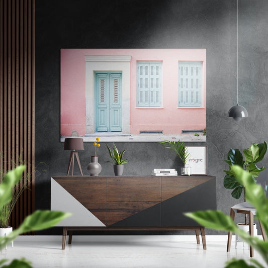 Brushed aluminium Dibond wall art displaying pink-walled building with turquoise door and white shutters, hung above wooden console table in modern interior.