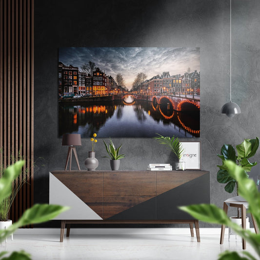 Brushed aluminium Dibond wall art depicting illuminated Amsterdam canal with bridges and historic buildings at dusk, displayed above wooden console table in modern interior.