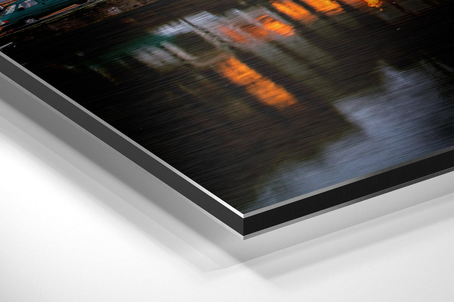 Close-up angled view of brushed aluminum Dibond wall art print with black border, showing layered edge construction and motion-blurred river landscape with orange and blue tones.