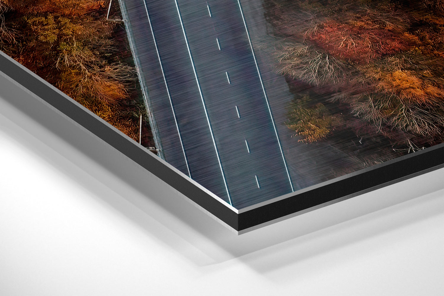 Angled perspective view of brushed aluminium Dibond wall art featuring an aerial road with lane markings and autumn trees, mounted on layered white and dark frame.