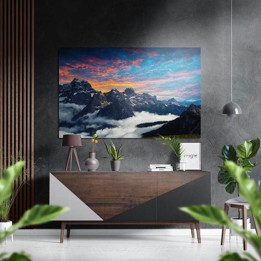 Brushed aluminium mountain landscape artwork hanging above wooden dresser in modern interior, displaying snow-covered peaks at sunset with orange and blue sky.