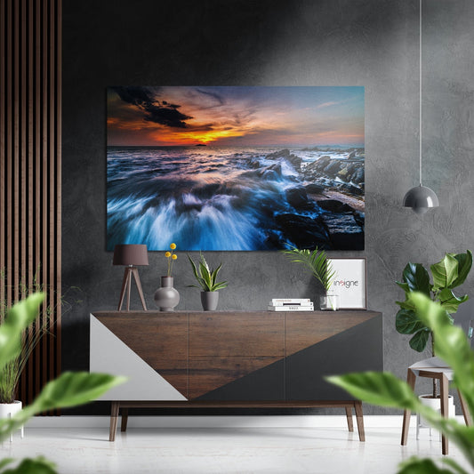 Brushed aluminium Dibond wall art displaying a coastal sunset with golden sky, blue waves, and rocky shore, displayed above a wooden sideboard in a modern interior.