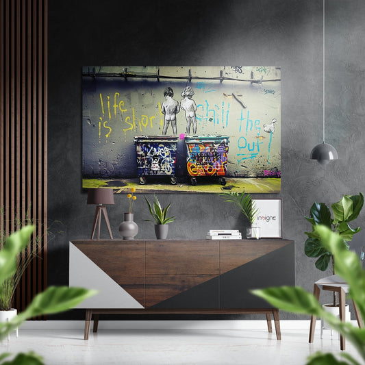 Street art aluminium wall panel depicting two nude figures above colorful graffitied dumpsters with yellow and blue text, displayed above a modern wooden console in a minimalist interior.