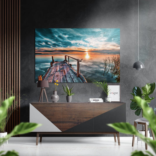Wooden pier extending into calm water at sunset with turquoise sky, orange clouds, and flying birds reflected on brushed aluminium panel.