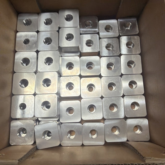 Fifty square metal clamp plates with centered holes arranged in cardboard box, overhead view.