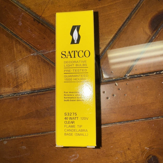 Yellow SATCO S3275 decorative light bulb box lying on wooden surface with metal pipe visible.